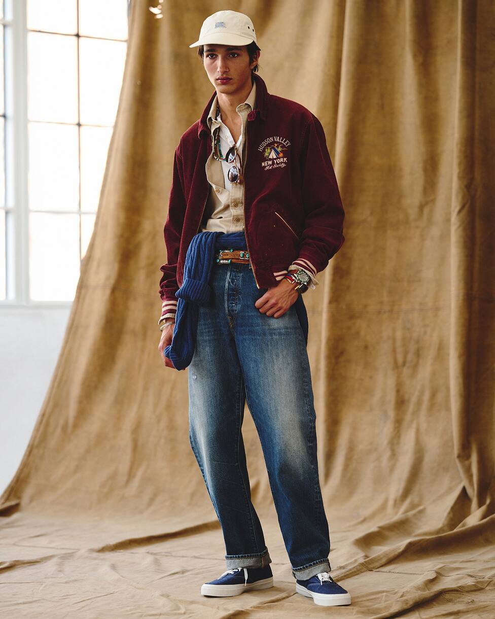 Man wearing a varsity jacket layered over a top and shirt, with jeans and a cap.
